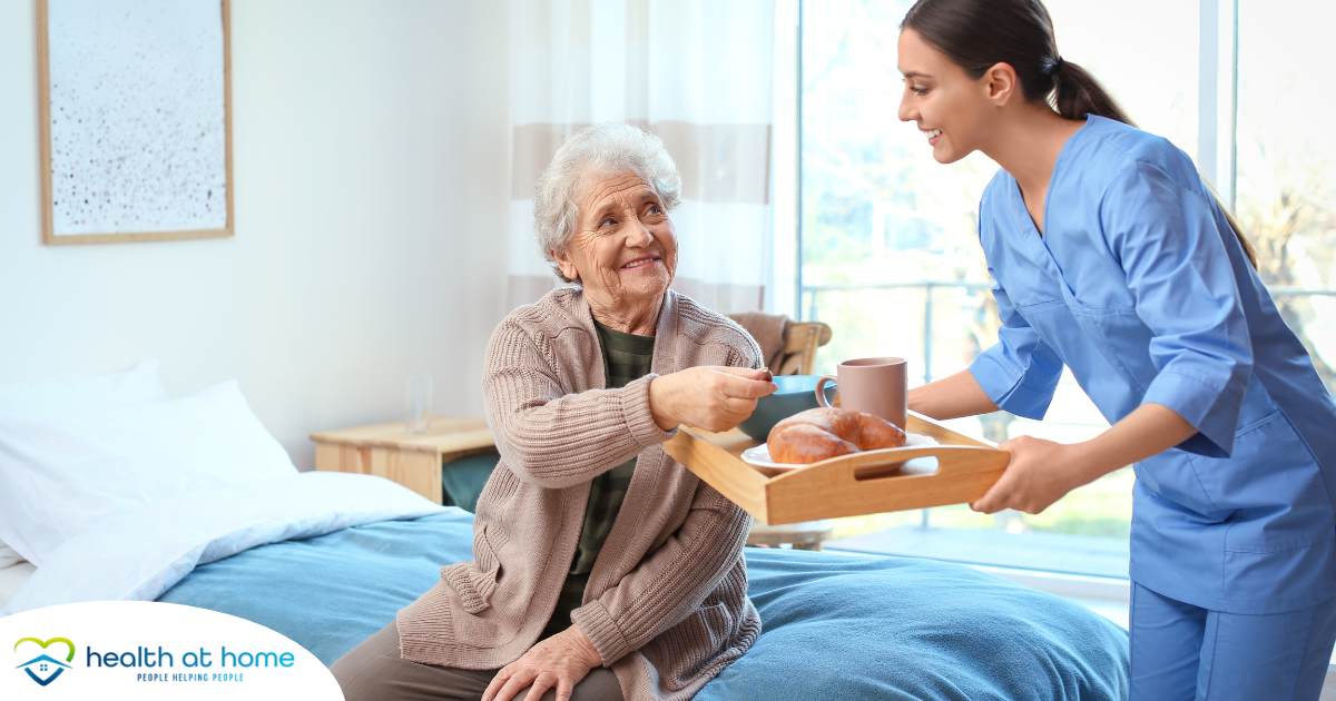 A professional caregiver smiles and provides food to an older adult in her care, representing the compassionate care needed for those with dementia.