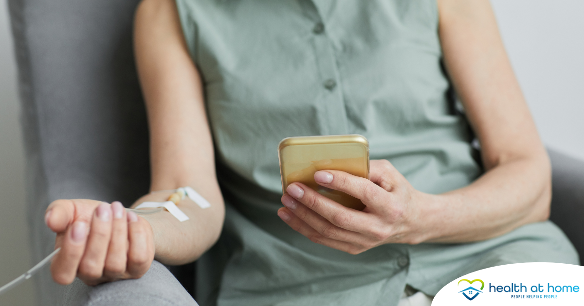 A woman comfortably receives IV therapy at home while using her phone.