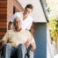 A caregiver brings an elderly man in a wheelchair out of a building, demonstrating a hospital discharge.