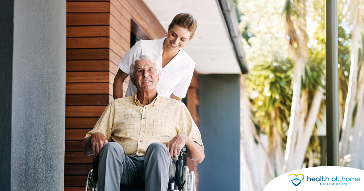 A caregiver brings an elderly man in a wheelchair out of a building, demonstrating a hospital discharge.